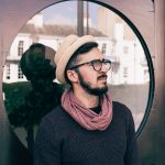 Portrait of a fashionable man wearing a hat and scarf, standing outdoors with a reflective background.